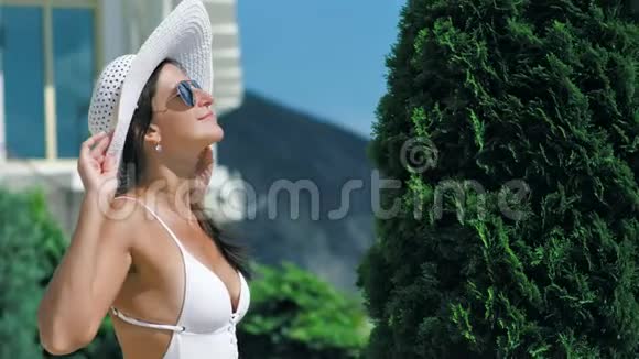戴着帽子和太阳镜的时尚女性在夏季酒店公园享受日光浴和微笑视频的预览图