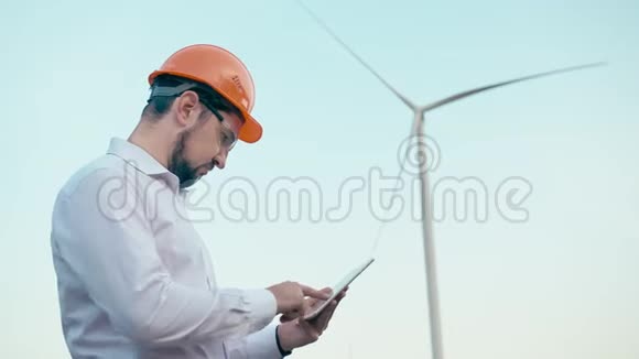 戴着橙色防护头盔和护目镜的高加索工程师在风力发电背景下使用平板电脑视频的预览图