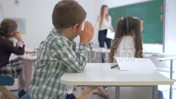 心烦意乱的小学生坐在教室里的桌子旁同学和女老师在黑板旁的背景下视频的预览图