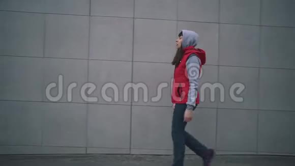 穿着红色无袖夹克的年轻女子走在大楼的灰色墙壁旁边视频的预览图