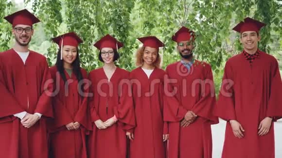 多民族毕业学生站在户外穿着红色长袍和灰泥板微笑和视频的预览图