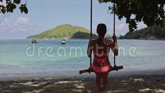 一个年轻的女孩穿着泳衣在自制的绳索秋千上摇摆在蓝色泻湖的沙滩上视频的预览图