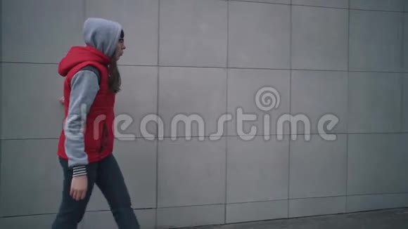 穿着红色无袖夹克的年轻女子走在大楼的灰色墙壁旁边视频的预览图