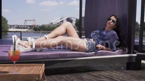 美丽快乐的女孩乌黑的长发和裸露的长腿在阳光下躺在休息室里戴墨镜的女人视频的预览图