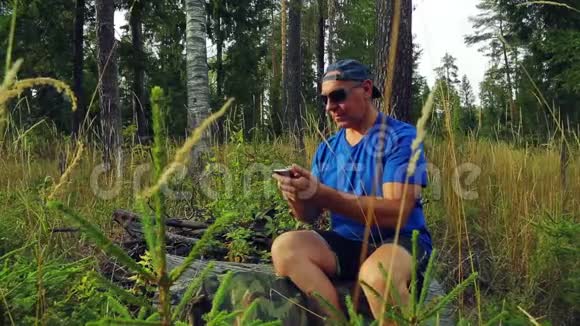 阳光明媚的一天一位戴着太阳镜和棒球帽的游客坐在一棵倒下的树上在森林里交流视频的预览图