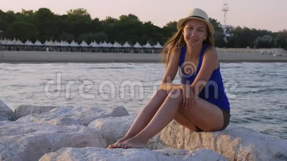 穿着蓝色T恤的快乐女孩巴拿马坐在海边的白色石头上慢动作视频的预览图