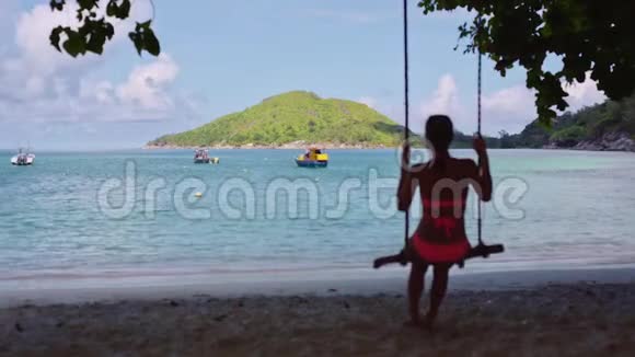 一个年轻的女孩穿着泳衣在自制的绳索秋千上摇摆在蓝色泻湖的沙滩上视频的预览图
