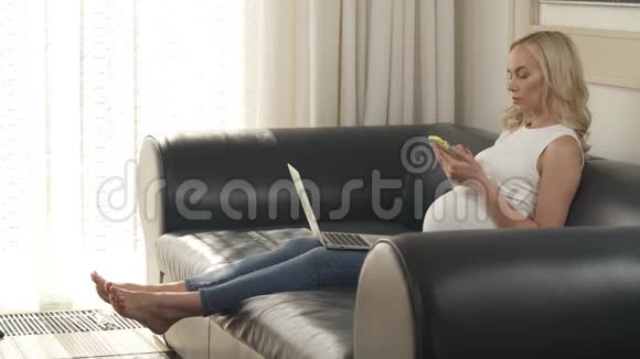 一个怀孕的金发女郎坐在沙发上拿着手机和笔记本电脑的侧视图她拿着手机视频的预览图