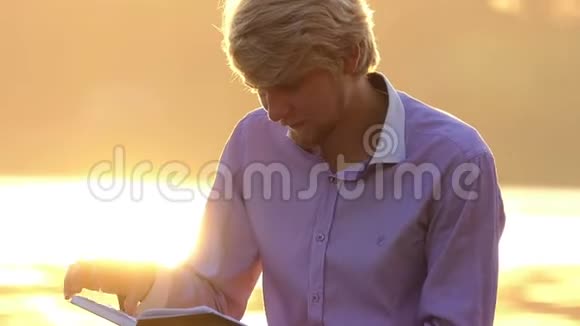 年轻的学生坐在湖岸上读一本手册视频的预览图