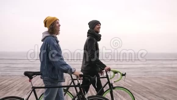 在秋日微笑的一对年轻的嬉皮士和他们的自行车一起在海边散步走过去视频的预览图