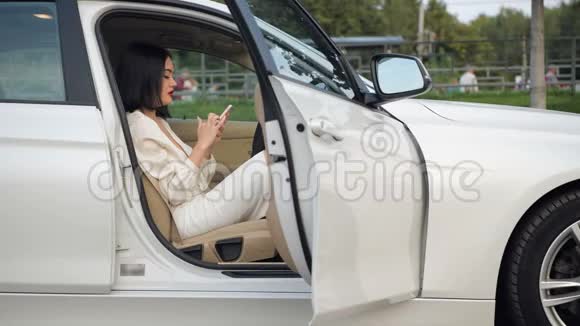 年轻漂亮优雅的黑发商务女司机在停车场通过电话交流在她的电话上输入一条信息视频的预览图