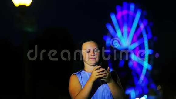 海滨的一位年轻女子在一个发光的摩天轮的背景下在手机上写了一条信息视频的预览图