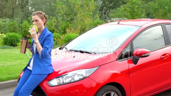 穿着蓝色西装的女人坐在红色汽车的引擎盖上吃午饭女孩吃三明治和热水瓶的饮料她很冷静视频的预览图