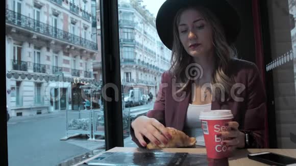 戴着帽子的漂亮法国女人在法国街头背景的咖啡店里吃羊角面包在桌子上视频的预览图