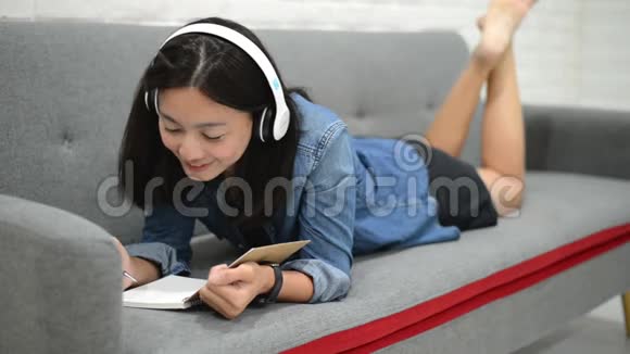 快乐的年轻女人躺在沙发上在笔记本上画一些东西听音乐视频的预览图