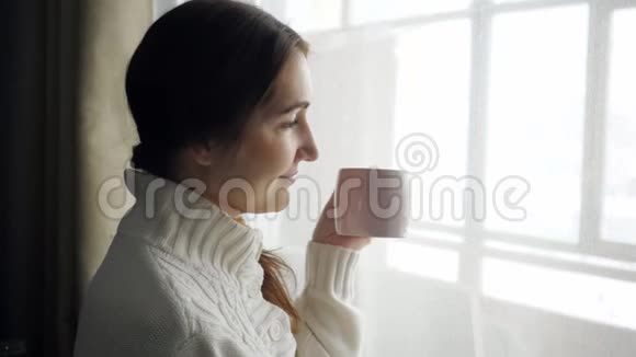 年轻漂亮的黑发女人穿着毛衣在窗户附近喝着杯咖啡视频的预览图