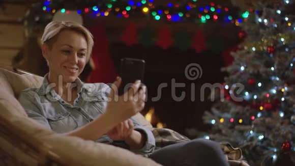 金发美女在圣诞夜壁炉前用智能手机视频聊天视频的预览图