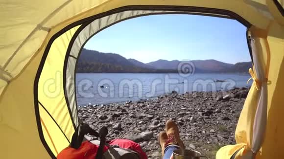 女旅行者躺在黄色的帐篷里女人手里拿着一杯茶山湖的景色然后视频的预览图