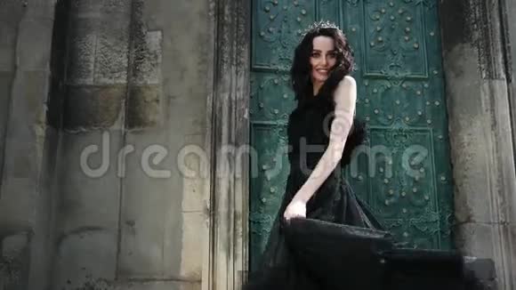 苗条的女孩黑发穿着黑色连衣裙戴着皇冠摆着姿势靠近老建筑视频的预览图