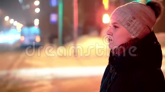 一个戴着冬帽的年轻女孩站在红绿灯前在夜光掠过的背景下视频的预览图