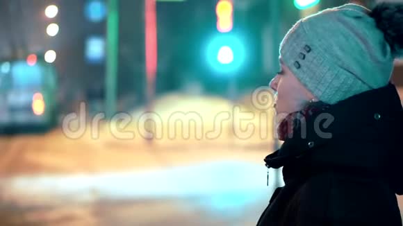 一个戴着冬帽的年轻女孩站在红绿灯前在夜光掠过的背景下视频的预览图