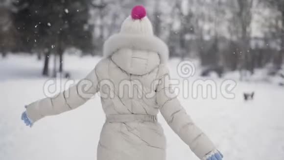 布鲁内特的头发是年轻的女人她的嘴唇是粉红色的穿着米色的羽绒大衣和帽子远离相机微笑着在雪中调情视频的预览图