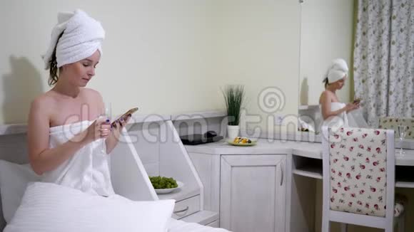 头上戴浴巾的女性饮酒在白色床上坐在移动互联网上视频的预览图
