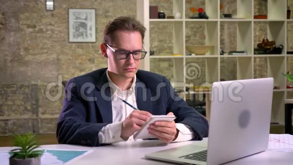 集中精力的白种人正在他的笔记本电脑前写笔记坐在灯光下的办公室里视频的预览图