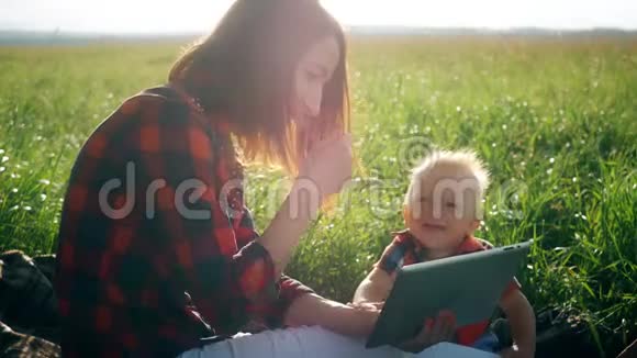 母亲带着婴儿在大自然中使用平板电脑幸福的家庭母亲和儿子带着平板电脑在公园里休息视频的预览图