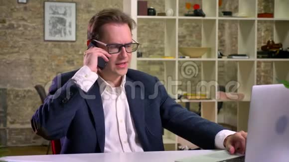 重要的电话和谈话白种人男性在办公室看笔记本电脑白桌砖墙视频的预览图