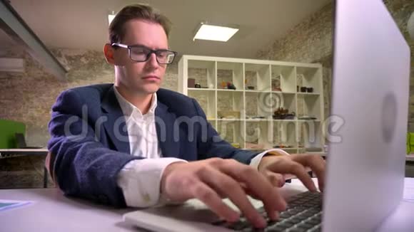 酷酷的白种人工作的男性戴着眼镜和西装专注于在笔记本电脑上打字独自坐在办公室里视频的预览图