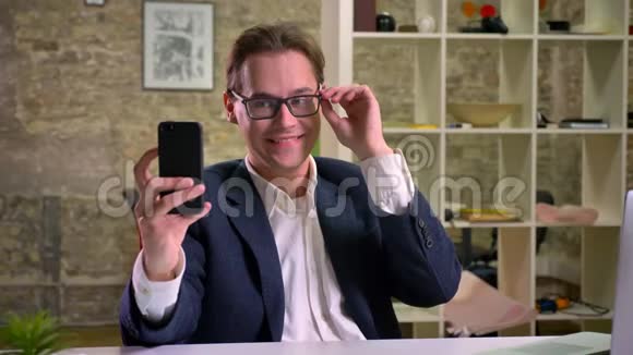 可爱的白种人男性西装革履一边在智能手机上自拍一边在办公室情绪视频的预览图