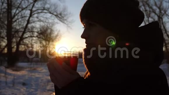 美丽的女孩在冬天的公园日落时喝着玻璃杯热茶在寒冷的空气中蒸汽从一杯视频的预览图