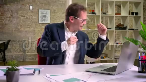 白种人男性坐在办公室里的笔记本电脑附近的超级有趣的舞蹈视频的预览图