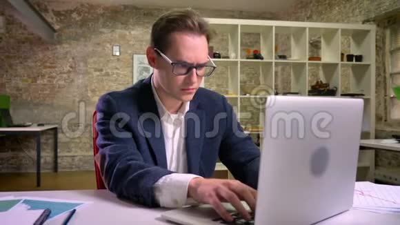 喜欢白种人的男人在室内笔记本电脑附近的工作场所展示胜利者的手势视频的预览图