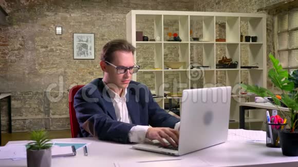 在办公桌前满意的白种人使用笔记本电脑而坐在后面的青砖视频的预览图