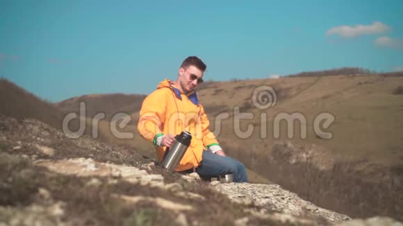 一个穿着黄色夹克蓝色牛仔裤和眼镜的男人坐在山上欣赏风景喝热水瓶里的茶视频的预览图