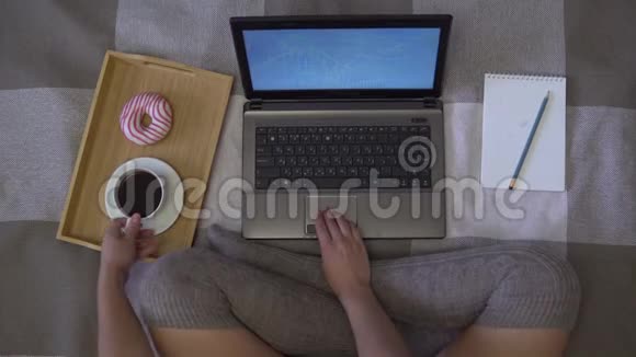一个女孩坐在床上用笔记本电脑工作用甜甜圈喝咖啡视频的预览图