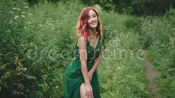 在镜头前摆姿势的野生森林里一个穿着绿色翡翠连衣裙的红色卷发女孩微笑轻轻地抚摸他视频的预览图