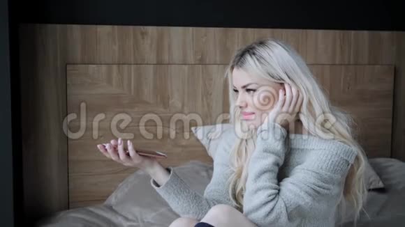 美丽的金发女人坐在床上用他的手自由地在电话里交谈智慧的概念视频的预览图