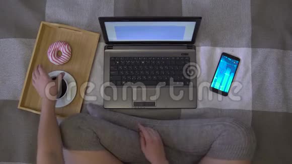 头顶射击女孩使用笔记本电脑坐在床上吃甜甜圈喝咖啡视频的预览图