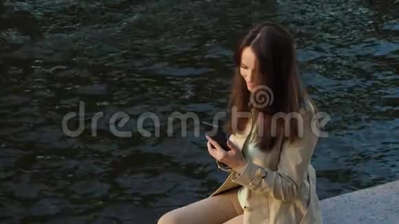 女人在智能手机上聊天坐在河边公园视频的预览图
