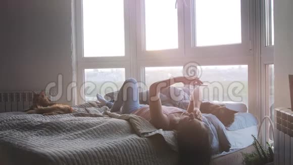 年轻的美女和她可爱的缅因州浣熊猫躺在床上放松在此期间她用手机靠窗视频的预览图
