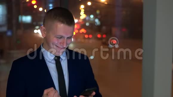 男商人在智能手机上读好消息微笑着靠近窗户与夜城慢动作视频的预览图