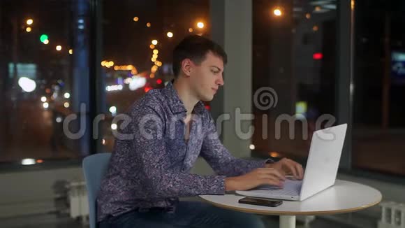 一个男人在笔记本电脑键盘上打字在办公室的一个大窗户里对着夜城视频的预览图