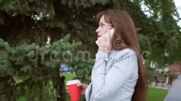 性格开朗的商务女性戴着浅色西装上班女孩在公园喝咖啡在智能手机上讲话视频的预览图