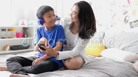 母亲和儿子戴着耳机用智能手机在家里一起唱歌视频的预览图