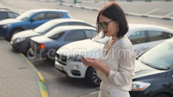 快乐的年轻女子穿着白色衬衫用手机在马路附近的街道上微笑在欧洲城市的汽车视频的预览图