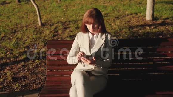 美丽的女商人戴着眼镜坐在长凳上和平板电脑打交道查看夏日公园里的电子邮件视频的预览图