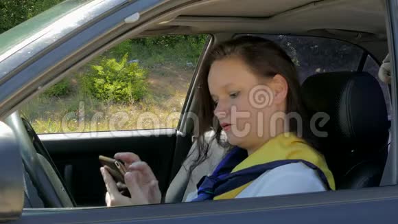 女人坐在汽车车轮后面使用智能手机视频的预览图